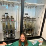 Kate Lucas poses by two racks of sediment cores in a large incubator. on July 2, 2024